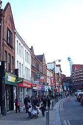 A row of shops facing onto a very busy pavement, with a large six-storey brick building visible in the background