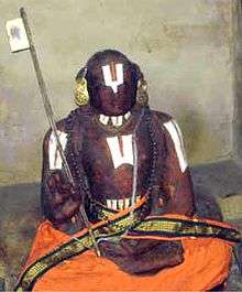An idol made of black stone dressed in orange robes, with a flag in a hand and Thirunamam on the forehead, throat, chest and hands.