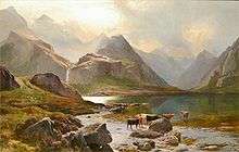 Tall, rocky mountains tower over a small lake, beyond which a waterfall cascades down from the heights. Brown and black cattle stand by the lakes margins, lit by wan sunlight that streams through the clouds.