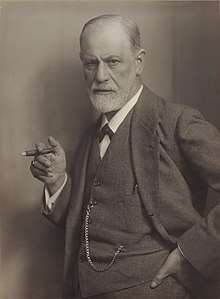 photograph of a balding man with a beard; holding a cigar and wearing a suit