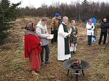 Eight people, all white, stand on heathland. Some of them are dressed in historical clothing akin to that worn in the medieval period.