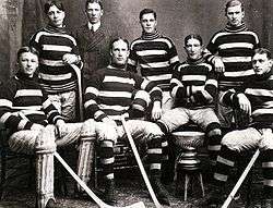 Seven men in hockey uniforms, four sitting in the front row, around the Stanley Cup trophy. Three stand in the back, with a man in a suit standing behind them.