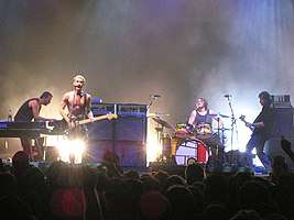 A rock band, Silverchair, performing onstage. From left to right, keyboardist playing a synthesizer, a drummer playing drumkit, a singer with short blonde hair and no shirt (also playing electric guitar), and a bass guitarist.
