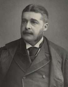 Upper body shot of a young man in a suit, with short hair parted in the middle, moustache and sideburns.