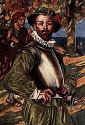 A bearded man, wearing green shirt and doublons, stands with his hands on his hips. The sea is visible in the background. A cuirass (metal armor) protects his upper torso. He is also wearing a green hat with a red rose and a white ruff on his neck. A rapier hangs ready at his left hip. Behind him on his right, a man is walking by, carrying a laden rucksack over his shoulder.