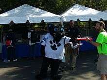  An anthropomorphic black cat, wearing a loose football jersey, is standing in front of several tents and is handing an object to another person.