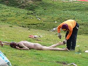 A dead, naked adult male body lies face down on a grassy area. Another individual, fully clothed and wearing gloves, has a knife and is cutting away part of the deceased body's foot. Other remnants of flesh are scattered about in the background.