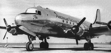 Close-up, in black-and-white, of a silver-bodied four-engined propeller aircraft standing on an apron at an airport