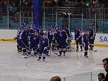 A group of ice hockey players skate on ice. They are wearing blue hockey jerseys and blue pants.