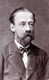 Portrait of balding, bearded, bespectacled middle-aged man with solemn expression, wearing a bow tie and high-buttoned jacket