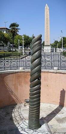 Photograph of a bronze spiral column, with an obelisk in the background