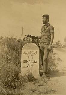 A bearded soldier with an Uzi next to a road sign specifying the distance to Ismailia