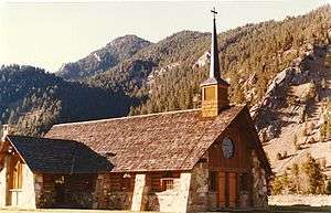 A stone church, seen from a corner and in the mountains with a short steeple and stained glass in the front.