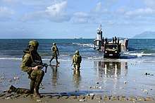 Colour photograph of three men wearing green military uniforms kneeling on a beach near a grey ship. A green truck is driving off the ship, and a large grey ship is visible on the horizon.