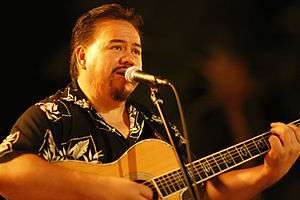 A man wearing a printed shirt, holding a guitar and standing behind a microphone