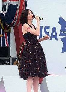 A British woman stands on a stage, wearing a black dress and holding a microphone in her left hand.