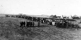 A slightly side-on front view of a line of early aircraft on a flat piece of land. People are standing with the aircraft, with a group standing in front of the machines. Several tent-like structures can be seen in the background.