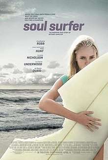 A young girl holds a surfboard at the beach. A section of her board is missing as if been bitten by a shark