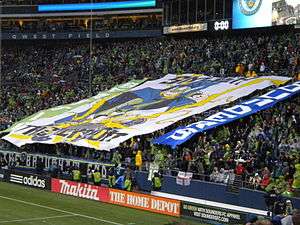 Fans in a stadium display a large banner. In the center is a picture of Thor wearing a green jersey and smashing the Union logo. It reads "Smash the Union" in the center, "Seattle" vertically on the left, and "Sounders" on the right.