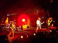 A rock band, Soundgarden, performing onstage. From left to right, a drummer playing drumkit, a guitarist (Kim Thayil), a singer also playing guitar, and an electric bassist. There are large, tall speaker cabinets onstage for the electric guitars and bass guitar.