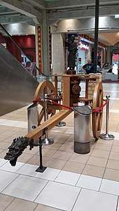 A wooden two-wheeled cart supporting a series of wooden peg gears and a metal statue pointing with one outstretched hand.