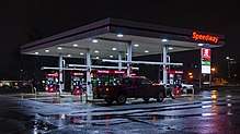 A white square canopy, seen from an angle with red gas pumps underneath, at night. At the one closest to the viewer, a red pickup truck is parked. On the canopy are illuminated red letters spelling out "Speedway"; the same word is also visible on the pumps and a sign behind the canopy where gas prices are given in green and red digital type. It is raining and the wet pavement reflects the lighted objects above it.