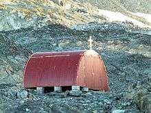 A small red Quonset Hut sittign on a concrete and metal pilings and topped with a cross sits among grey rocks.