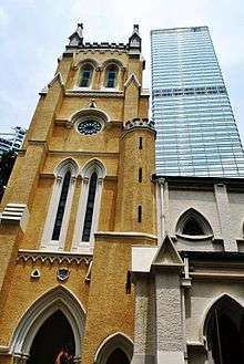 A pared-back Gothic-style yellow church tower seen from below, with a glass-surfaced skyscraper behind it.