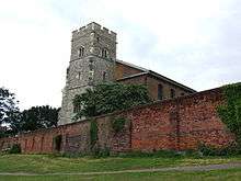 A grey stone tower rises above an ancient red brick boundary wall
