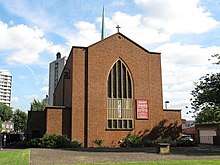 Tall rectangular brick church with arced stained glass window with external bell and copper spire.