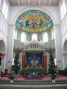 The sanctuary of a cathedral. On a raised dais, the low altar stands, flanked by Christmas trees. Behind it, the high altar is visible, with the tabernacle on it. A ciborium is over the high altar, directly under an arch with a domed apse behind it.
