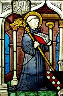 Stained glass image of a kneeling man with a halo holding an open book and a staff