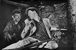 Aleksei Stakhanov and another man at work in a Soviet coal mine. Stakhanov, while holding a drill, is seated at the coal face, his head turned to speak to his colleague.