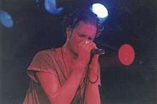 A male singer, Layne Staley, singing into a microphone at a concert. His eyes are closed as he sings. He is wearing rings on his left hand.