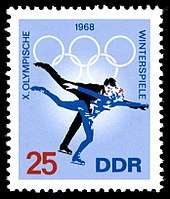 A postage stamp with a blue background and two figure skaters skating, the date 1968 is centered on the top of the stamp along with the Olympic rings. The word "Winterspiele" is written down the right side, the words "X Olympische" are written down the left side. The number 25 is in the lower left corner and the letters "DDR" are in the lower right corner