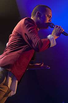 A man with brown skin wearing a red suit jacket and holding a microphone.