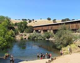 People at a river bank with a covered bridge in the background.