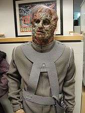 A mannequin wearing a face mask that resembles decomposing flesh and a grey body suit.