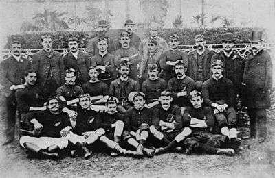 Photo of team players and management all of whom are seated or standing, in four rows, wearing either their playing jerseys with caps, or formal wear.
