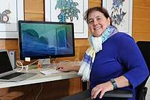 Stefi Baum at her desk wearing a blue shirt, scarf and there is a computer behind her.