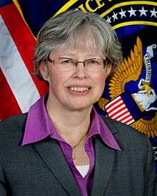 image of Stephanie O'Sullivan, smiling at the camera, wearing a collared purple top and dark coat, with U.S. and DOJ flags to each side in the background