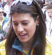 Front on image of a smiling, brown haired young woman with hair longer than her shoulders, pearl spherical earrings with sunglasses on her hair which is parted down the middle, wearing a yellow jacket tracksuit with a green collar, with a red medal ribbon around her neck.