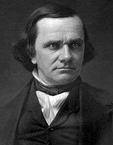 Photograph of a man with longish dark hair; he is wearing formal attire which consists of a dark vest, a white shirt, and a tie of the style worn in 1860.
