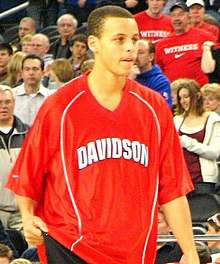 A young man in his early 20s is warming up for a basketball game. He is wearing a red warm-up jersey with "Davidson" written in white across the chest. He is a light-skinned male of mixed race (half white by his mother, half African American by his father).