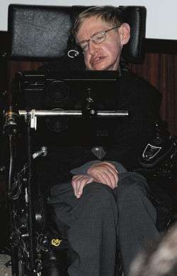 A colored photograph of a man, sitting in an wheelchair with his arms folded. He is wearing glasses and is looking from left to right.