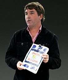 A photograph of a middle-aged man in a jacket standing and holding a book