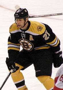 A hockey player in a Black uniform with gold trim and a stylized "B" logo on his chest defends his position during a game.