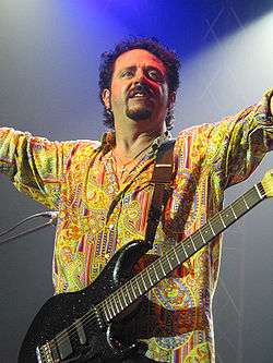 A man in a multi-colored dress shirt with a black guitar strapped around his neck.
