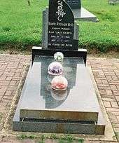 A grave and headstone. The structure is made from a shiny grey rock, and lacks ornamentation. Three flowers encased in circular glass baubles sit on top of it.