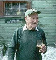 Color picture; An elderly man holding a glass and wearing a hat stands in front of a wooden lodge